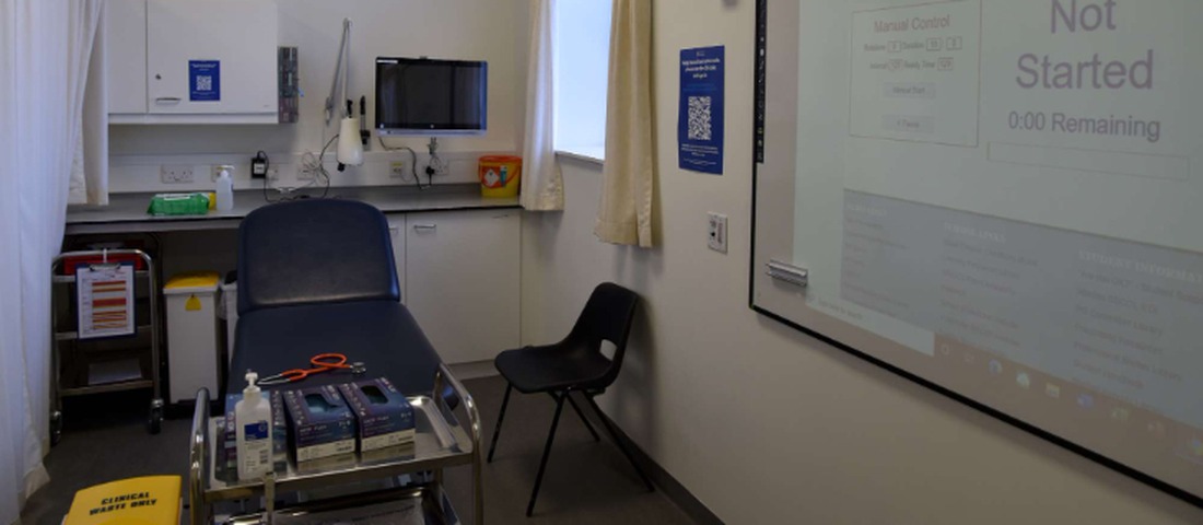 2021-04-22-OSCE-Set-Up-26-2000_1100x480 School of Medicine building with large windows and a wooden deck, bordered by a glass railing. A tree stands near the building under a clear blue sky