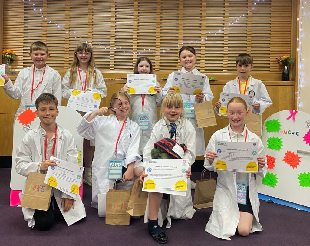 A group of primary school-aged children hold certificates to celebrate their participation in a research project to understand food and fairness from their perspectives. The children are wearing lab coats and have various props with them.