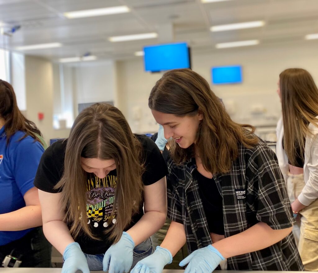 Two teenage female students wearing blue rubber gloves lean over a table inspecting something on it.